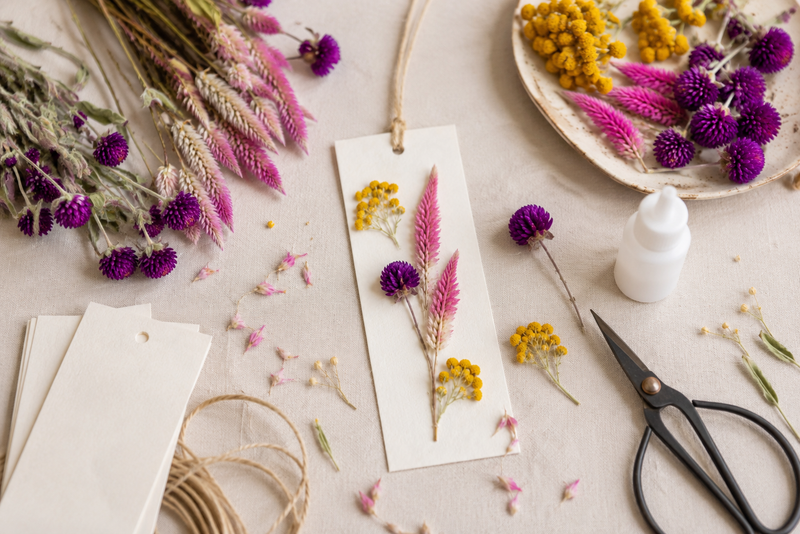Création d'un marque-page en fleurs séchées avec achillée jaune, célosie Flamingo, gomphrena violette, colle et ciseaux.
